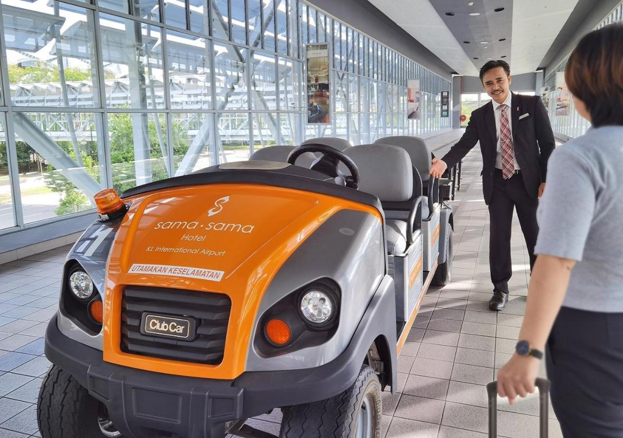 萨马萨玛克利亚酒店 雪邦 外观 照片 The photo shows a small electric vehicle, typically used for transportation in areas like airports or large venues. It is orange and black, with branding that includes the name "sama-sama." In the foreground, a man in formal attire stands next to the