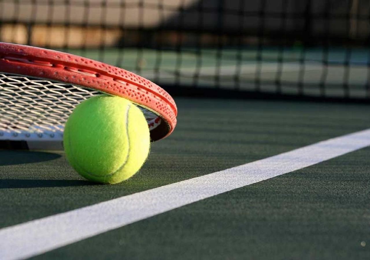 萨马萨玛克利亚酒店 雪邦 外观 照片 The photo shows a close-up of a tennis court. In the foreground, there is a bright green tennis ball resting on the court surface, next to a tennis racket that is placed against the net. The racket has a red frame and a white string pattern, and the 