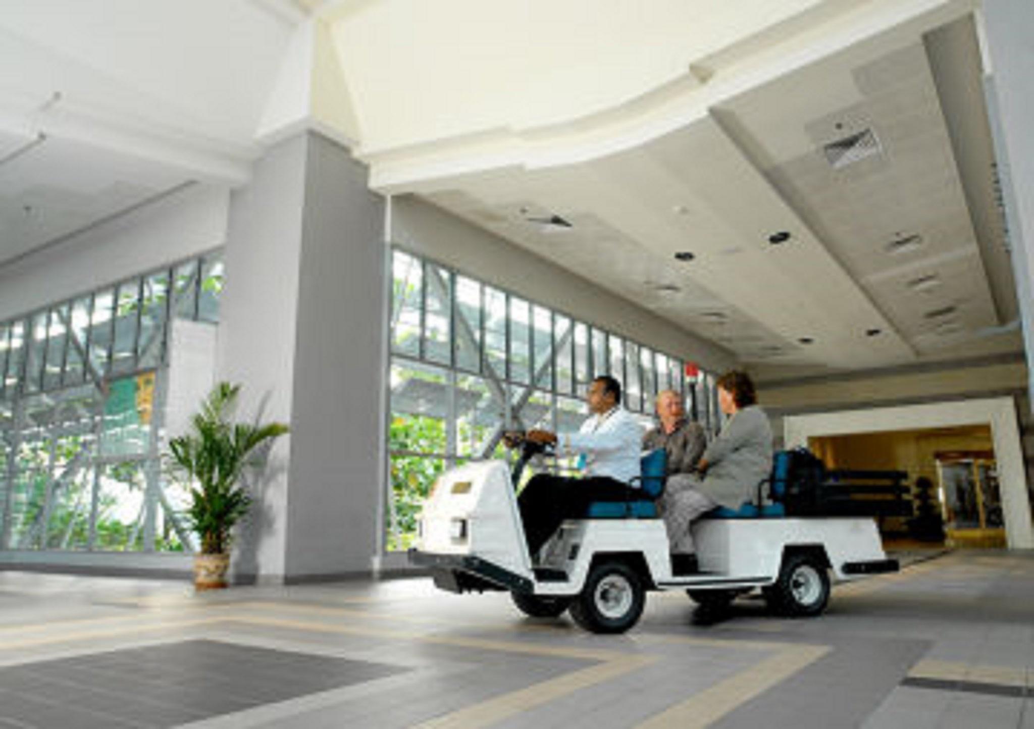 萨马萨玛克利亚酒店 雪邦 内观 照片 The photo shows a small vehicle, likely an electric golf cart or utility cart, driving inside a spacious indoor area. Three people are sitting in the cart, with two of them looking forward. The surrounding space features large windows allowing natura