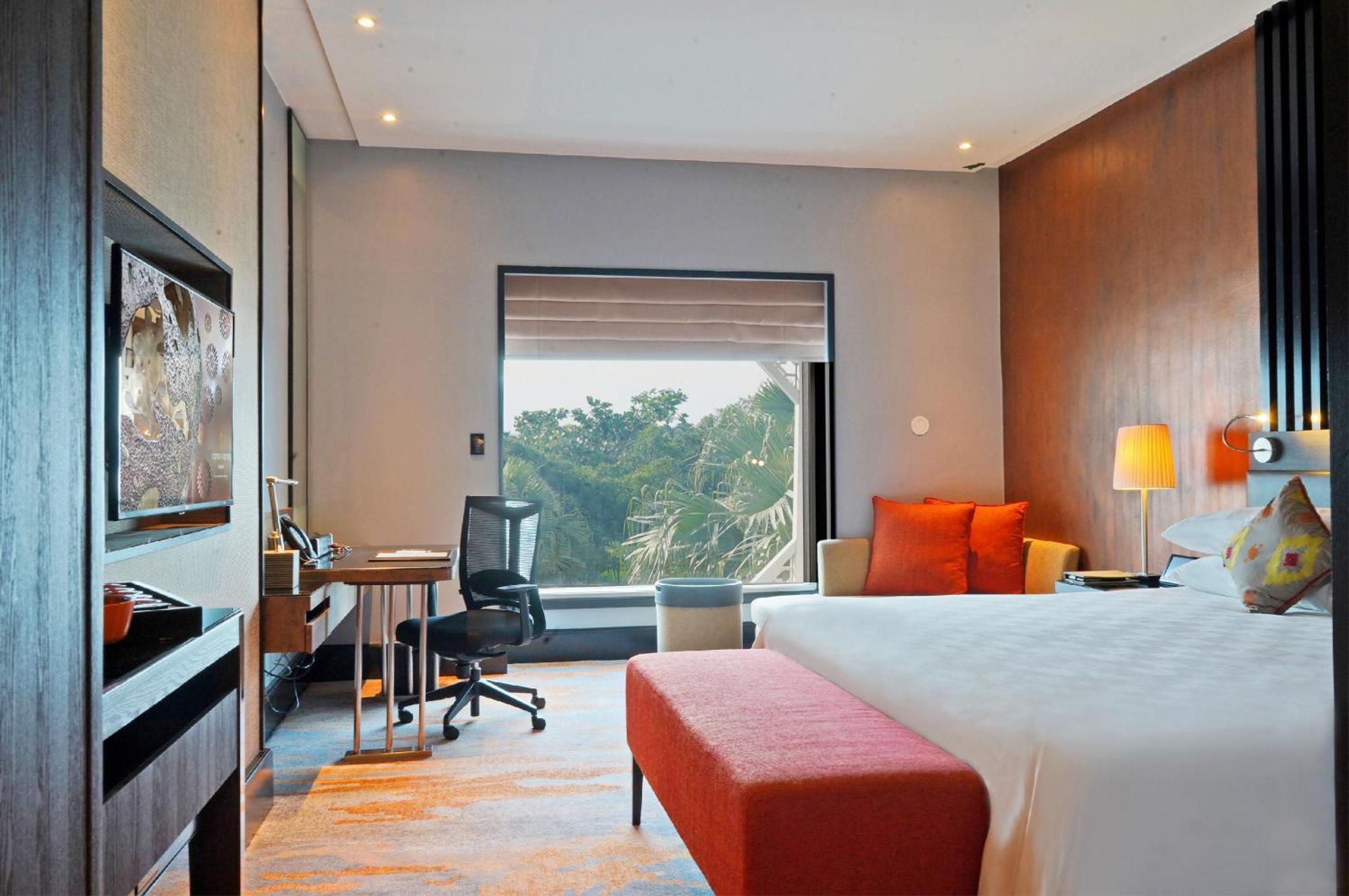 萨马萨玛克利亚酒店 雪邦 外观 照片 The photo shows a modern hotel room with a clean, contemporary design. There is a large bed with white linens and several orange accent pillows. A red upholstered bench is positioned at the foot of the bed. Beside the bed is a stylish chair and a sma